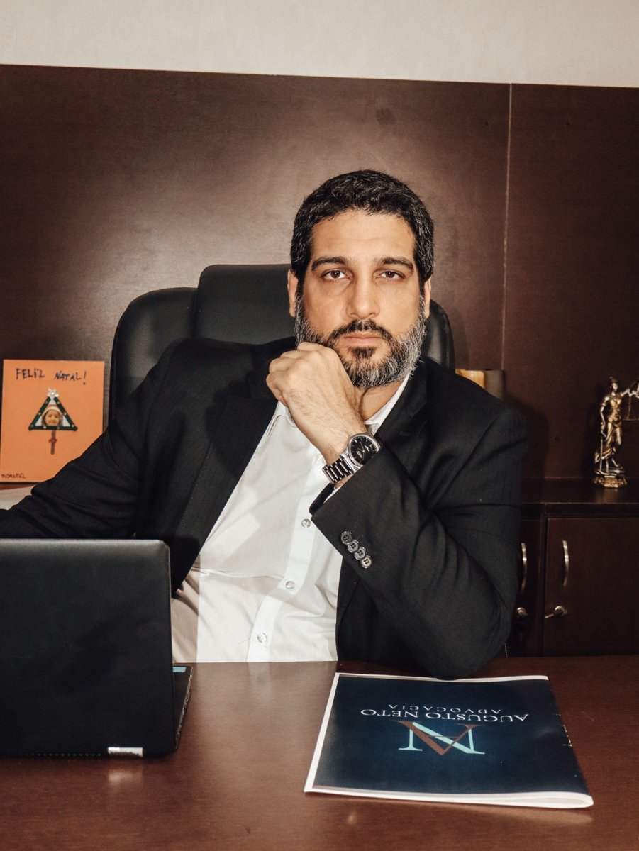 O advogado Augusto Neto sentado à mesa do escritório, de terno, com material da Augusto Neto Advocacia sobre a mesa